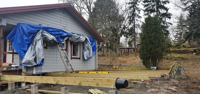 Hus under renovering med presenning, nya bärlinor på marken, och stolpskor samt grå himmel i bakgrunden.