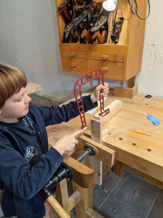 Barn arbetar med trä och verktyg vid en snickarbänk, håller i en röd mall för snickeriprojekt.