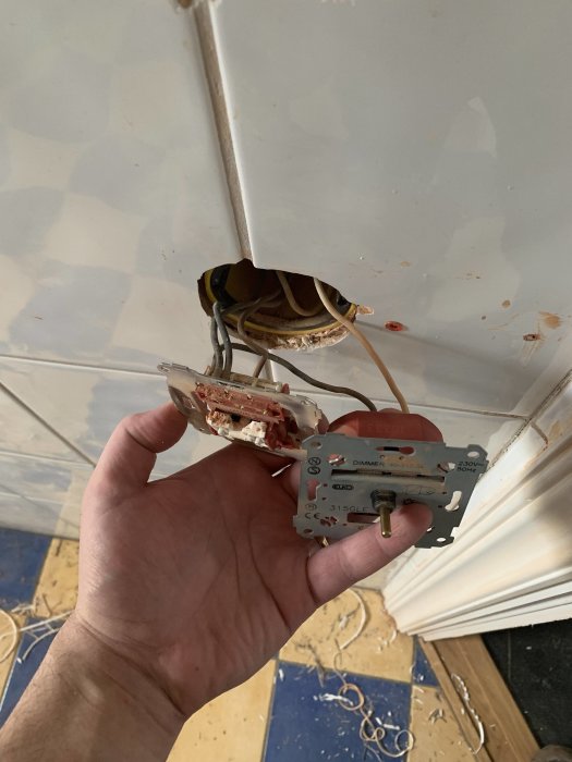 Hand håller gamla eluttag för infälld lampknapp och utanpåliggande dimmer mot vit vägg.