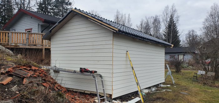 Färdigspikad panel och knutbrädor på ett uthusets kortsida, med spikpistol och stege framför på gräsmattan.