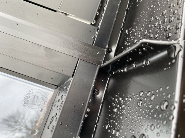 Närbild på svart fönsterbleck med vattendroppar, felaktigt monterad med synlig skruv och ojämn tätningsfog.
