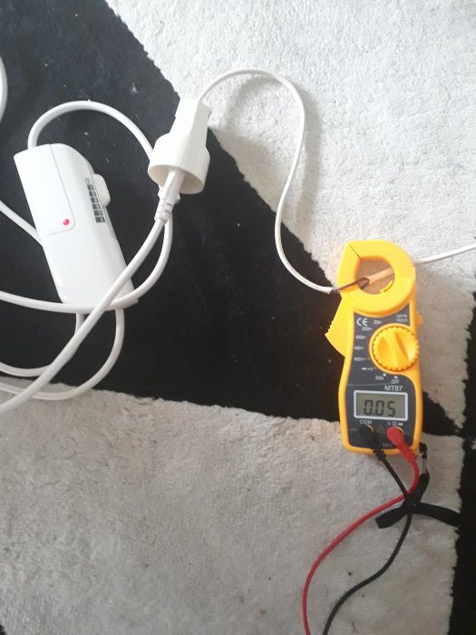 Tångmultimeter mäter ström på en fleecefilt med värmeslingor på ett mattgolv.