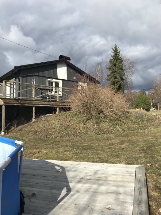 Bakgård med befintlig liten trädäck och gräshöjd framför svart hus, molnig himmel.