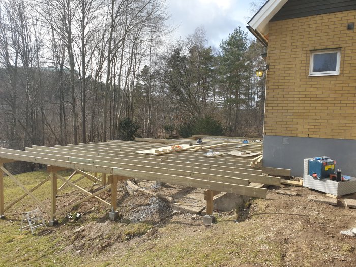 Pågående konstruktion av en stor träaltan med bärlinor och skruvreglar vid ett hus.