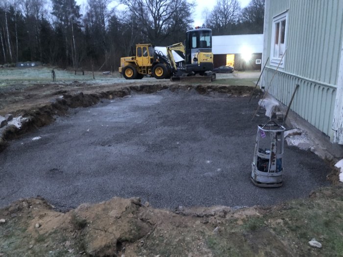 Grund med makadam, en padda framför, en minigrävare och en traktor i bakgrunden vid skymning.