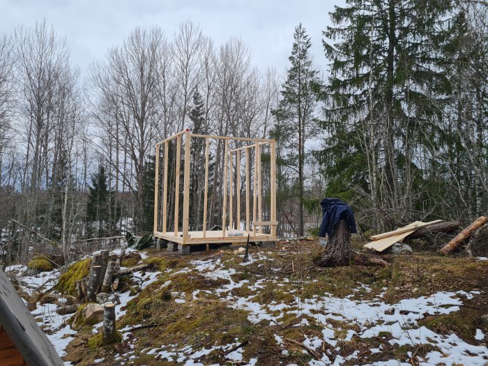 Stomme av ett trähus under konstruktion i en skogsmiljö med snöfläckar på marken.