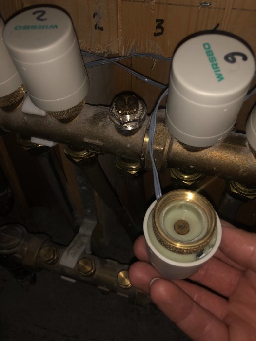 Hand som håller ett avlägsnat termostatdon bredvid en pinnstyrd värmedistributionsfördelare med synliga rörkopplingar.