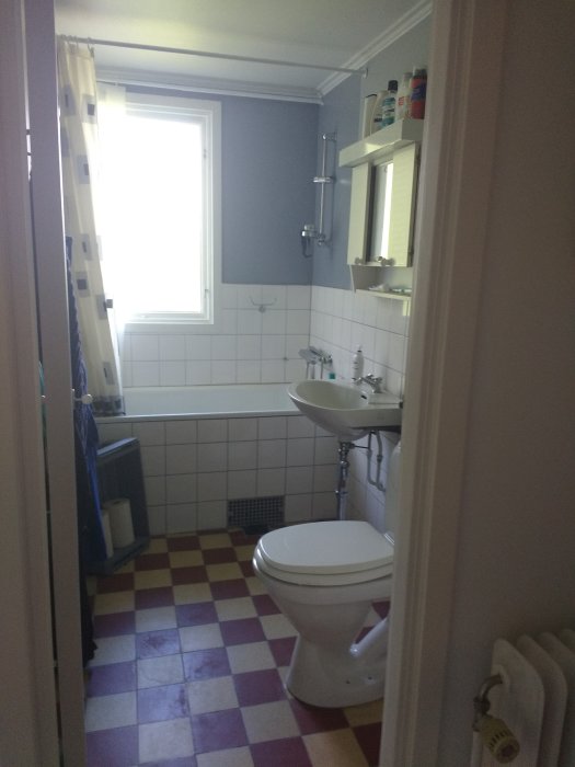 Badrum med vit WC och tvättställ, rutigt golv, badkar och fönster, före renovering.