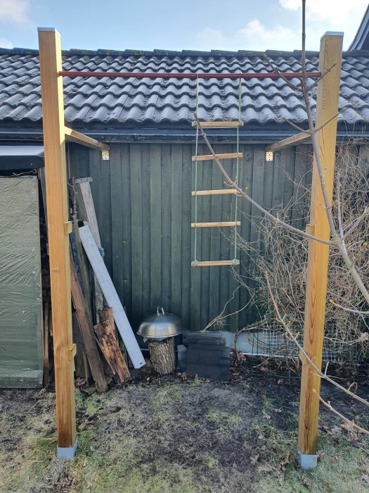 Nyuppställda trästolpar med ett rörmellertvärstag i en trädgård framför en stuga med tegelpannor och träavfall vid sidan.