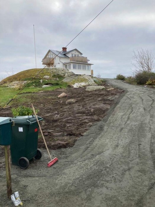 Hus på kulle med avloppsskorsten, grusväg i förgrunden, soptunna och röd kvast till vänster.