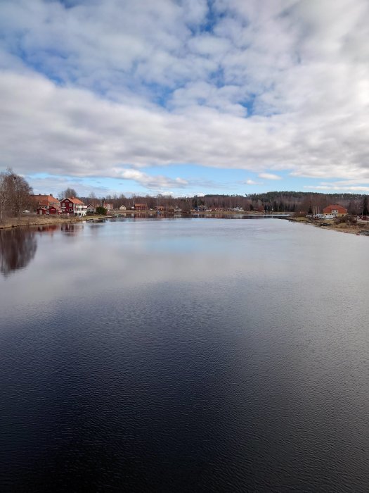 Vy över en lugn sjö med spegelblank yta och traditionella röda svenska hus längs strandkanten.
