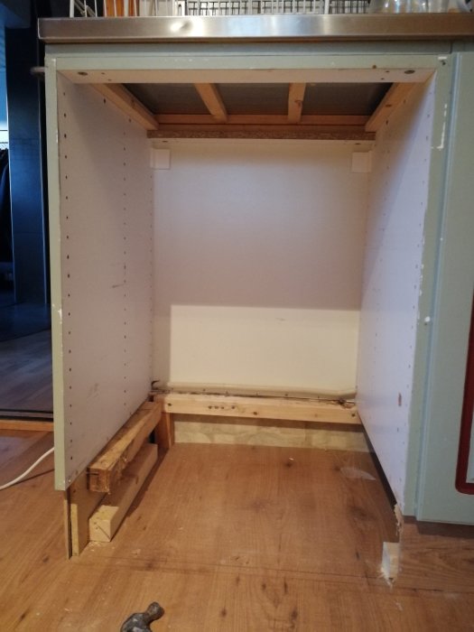 Utkapad plats i köksskåp med synliga reglar och spånskiva, förberedd för tillfällig diskmaskinsinstallation.