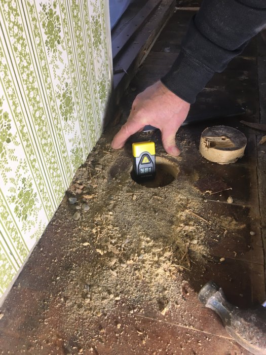 Person mäter fuktkvot i trägolv med digital fuktmätare nära hål med synlig sandunderlag.