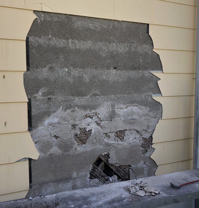 En delvis borttagen fasadbeklädnad avslöjar gammal puts och hönsnät på ett hus.