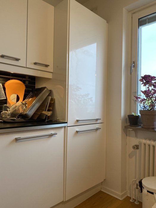 En vit köksinredning med en inbyggd kyl/frys vars front är repad, intill en diskbänk och fönster.