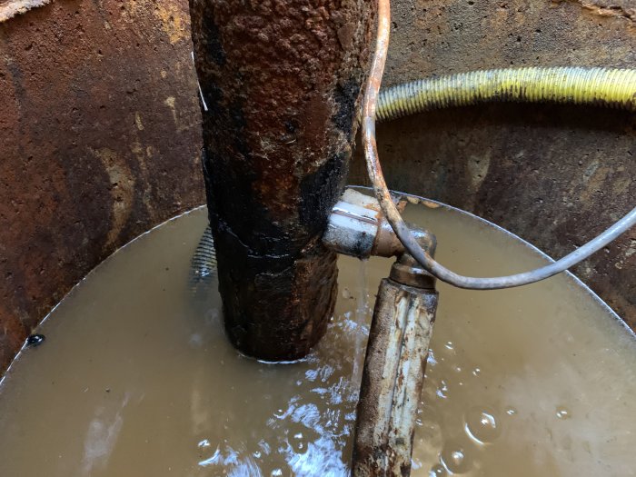 Brunn med lösa elkablar och kopplingsklämmor, järnrör tvärs över, vattensamling och vajer som antyder pump.