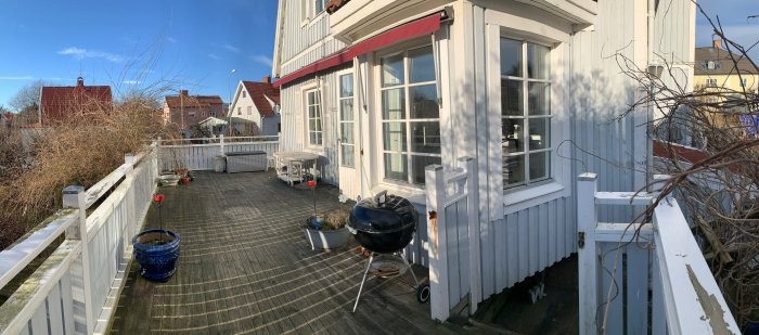 Trött altan vid ett hus med slitet trägolv, krukor, grill och vissna växter, redo för renovering.