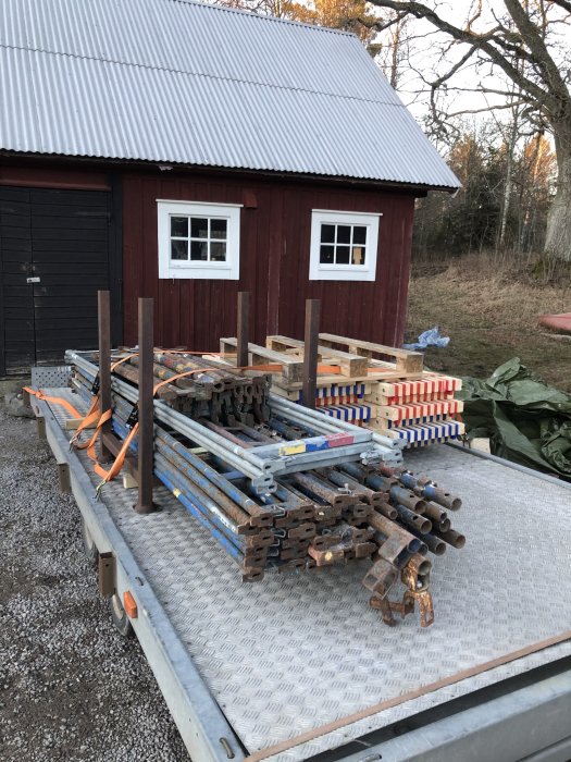 Haki-ställning delar lastade på släpvagn framför rött hus med plåttak.