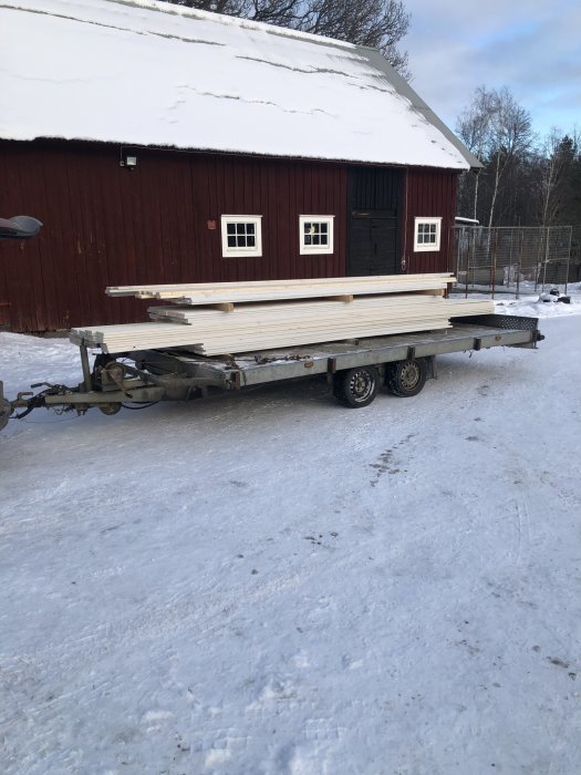 Släpvagn lastad med färdiggrundad panel framför röd byggnad på snöig mark.