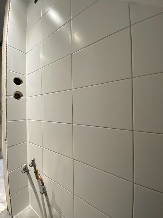 Nyligen fogade vita kakelplattor på en badrumsvägg med synliga rör och väntande anslutningar.