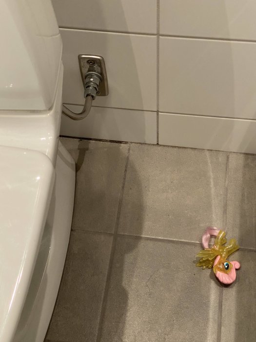 Golv och vägg i badrum med mörkare våt fog under handfatet och en plastfigur på golvet.