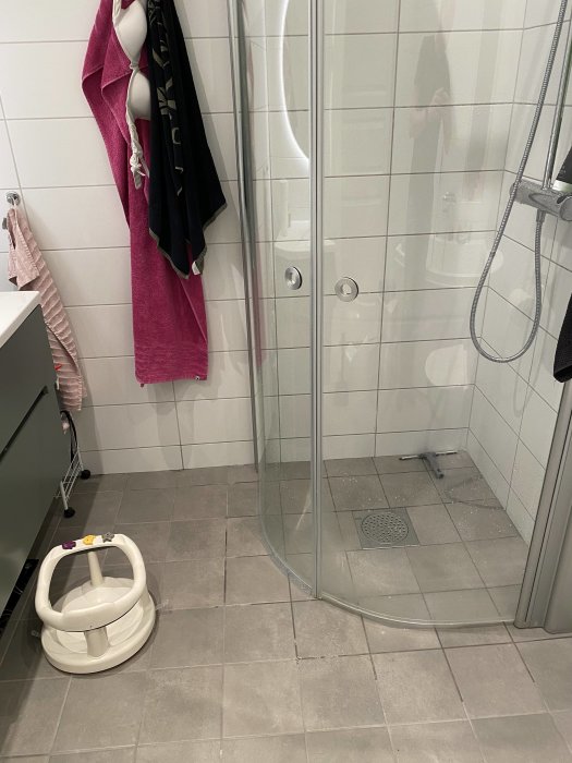 Badrum med duschhörna och grått kakel, vatten syns utanför duschen nära handfatets kant.