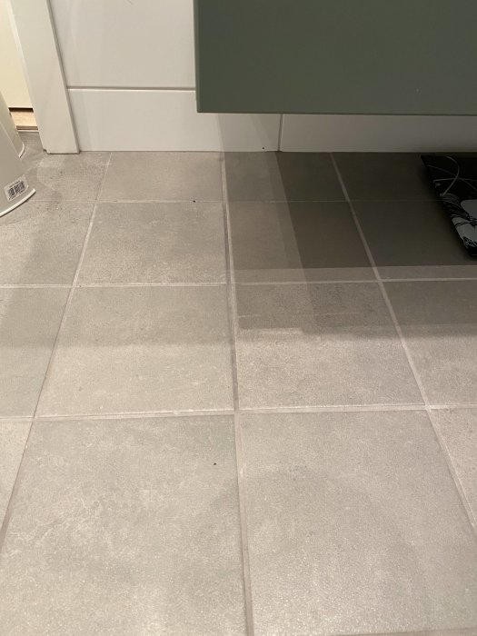 Gråa klinkergolv med fuktfläckar nära ett grönt skåp och vit sockel.