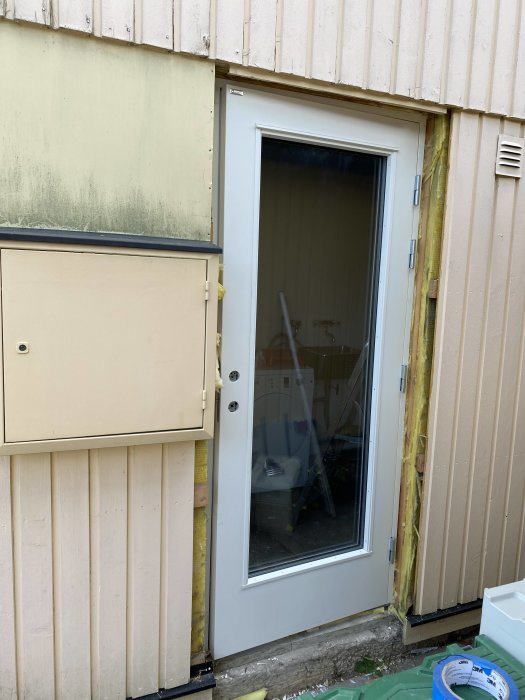 Husfasad under renovering med synlig gul isolering där ytterpanelen har tagits bort, bredvid en vit dörr.