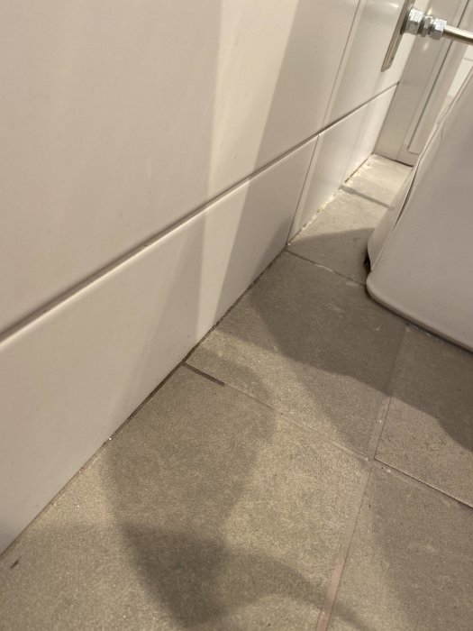 Hörn av ett badrum med vita kakelväggar och grått klinkergolv som behöver fogas om.