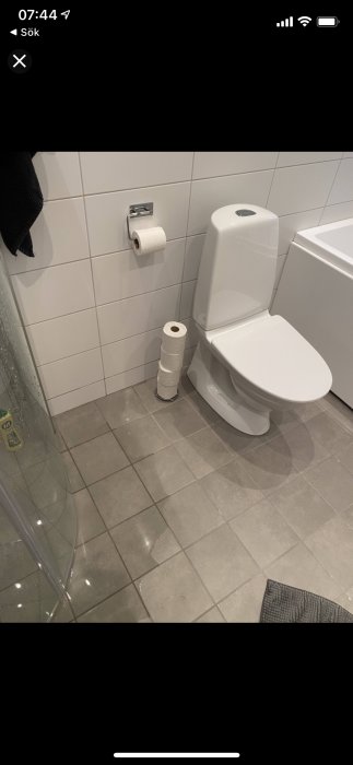 Toalett intill dusch med synliga vattendroppar på grått kakelgolv.