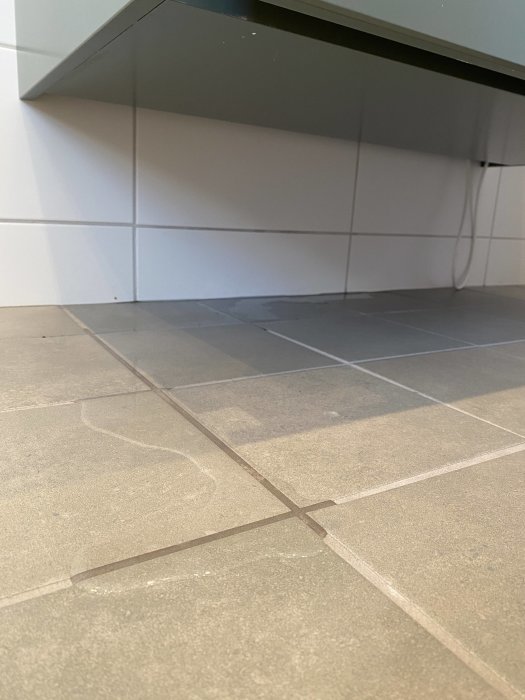 Vattenläckage på kakelgolv bakom badrumskommod nära vägg där rörledningar är installerade.