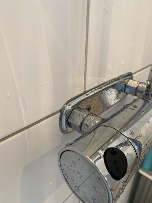 Närgräns av en silverfärgad duschblandare med vattendroppar på en vit kakelvägg i bakgrunden.