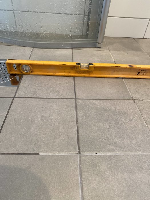 Vattenpass som ligger på klinkergolv intill en dusch med synlig lutning.