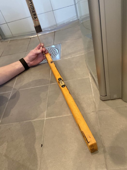 En hand håller en gul nivåsticka mot en lutande badrumsgolv nära en duschdränering för att kontrollera sluttningen.
