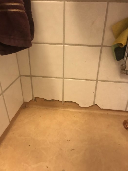 Skadat kakel i hörn nära golv med synliga brister längst kakelraden.