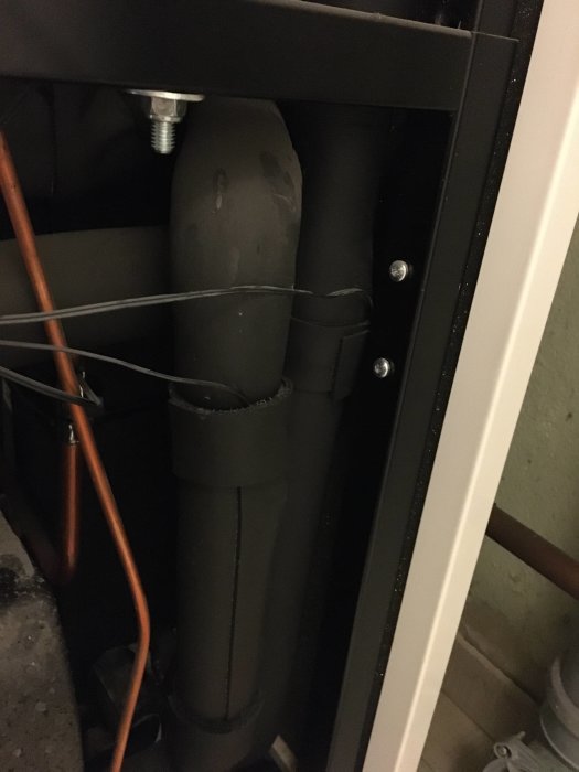 Svarta avloppsrör monterade vertikalt vid en vit dörrkarm, omgivna av kablar och rör.