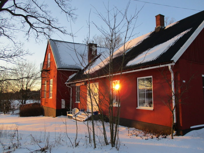 Röd husfasad med två delar under snötäckt tak i vintersolnedgång, den ena delen högre med solfångare.