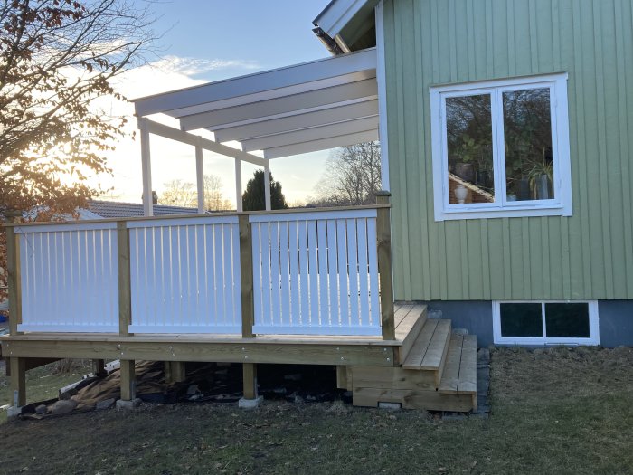 Ett nybyggt trädäck med vit räcke och överliggande tak med ca 240 cm höjd till dess främre bärlina, vid ett grönt hus.