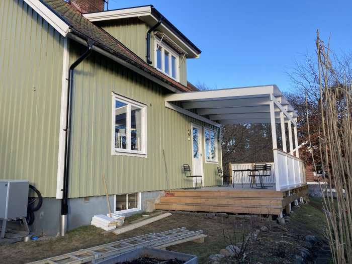 Upphöjd träaltan med högt satt tak nära ett tvåvåningshus, vilket ger ett luftigt intryck och öppnar upp utrymmet.