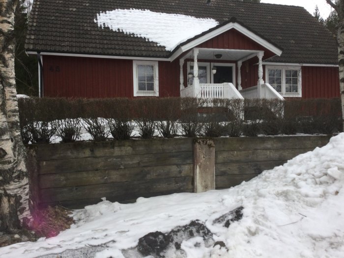 Ett rött hus med snötäckt tak och vit veranda bakom en stödmur i trä med snöhög framför.