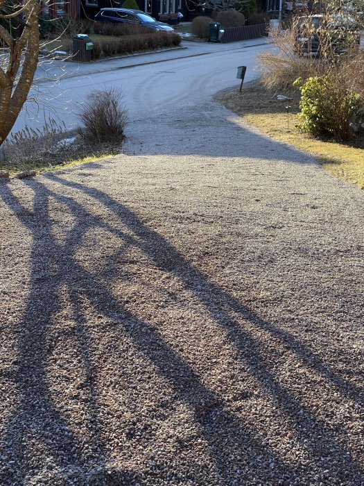 Grusad uppfart med varierande tjocklek av krossprodukt och skuggor av ett träd, i ett bostadsområde.