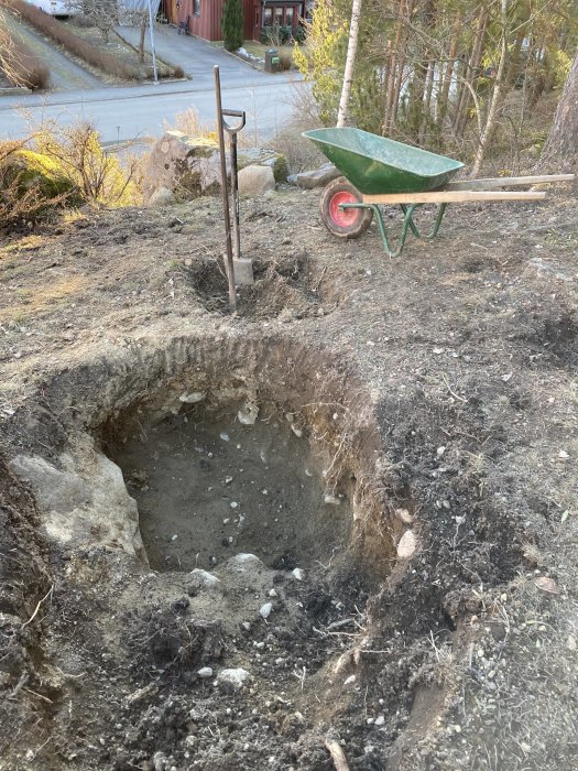 Nygrävd planteringsgrop för fruktträd med skottkärra och spade i bakgrunden, stenigt jordlager synligt.