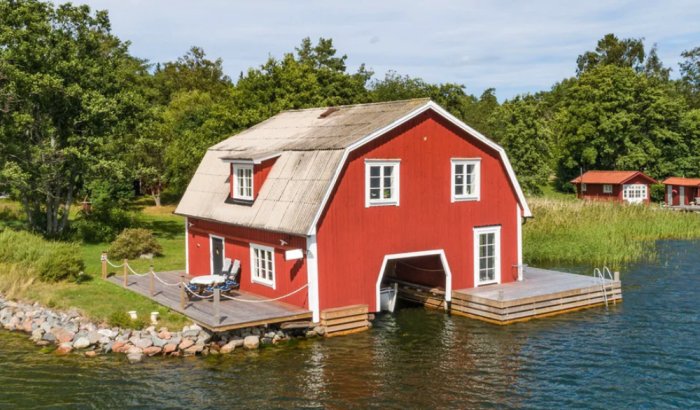 Röd sjöbod med öppning för båt, belägen vid vatten med brygga och omgivande grönska.