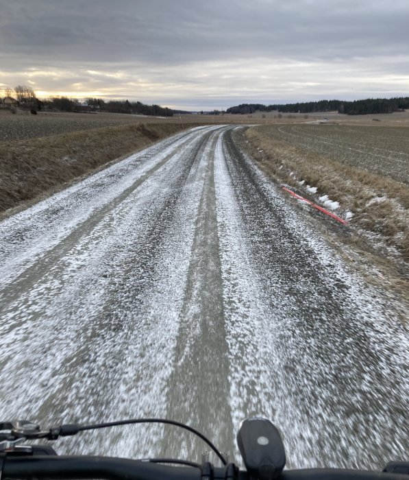 Vy från en fyrhjuling som färdas på en grusväg med lätt snötäcke, omgiven av öppna fält.