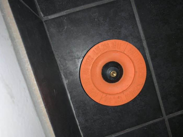 Svart kakelgolv med en orange täckplatta för rörgenomföring, relaterad till byggbesvär.