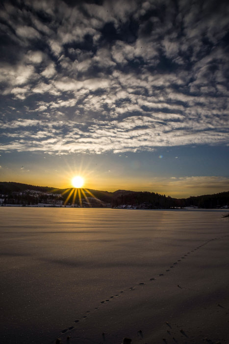 Solnedgång över en frusen sjö med fotspår i snön och moln på himlen.