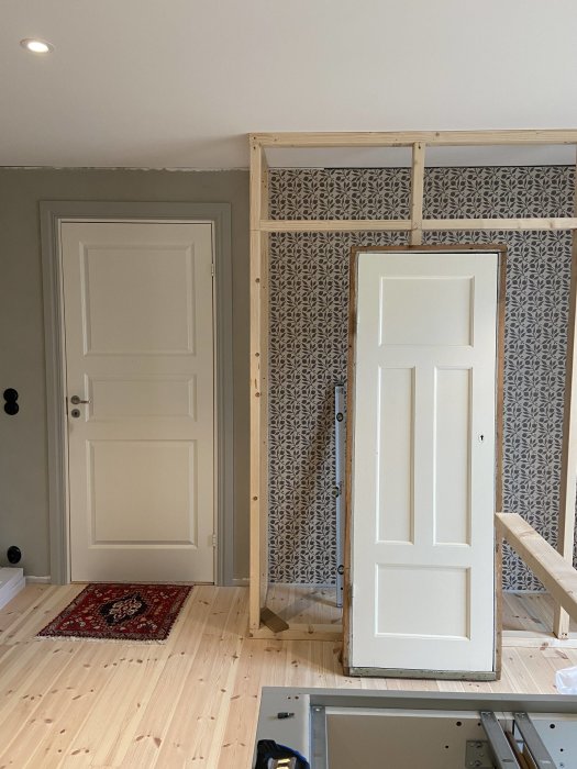 En pågående skafferibyggnation med trästomme och en vit dörr, inuti ett rum med tapeter och trägolv.