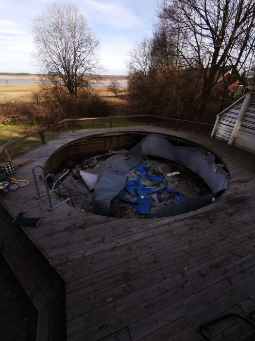 Sönderslagen rund pool med blå presenning på en träterrass, med sjöutsikt och träd i bakgrunden.