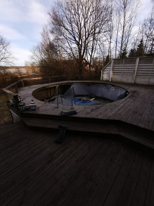 Delvis tömdt ovalt badkar på en stor träterrass omgiven av träd och staket med verktyg runtomkring.