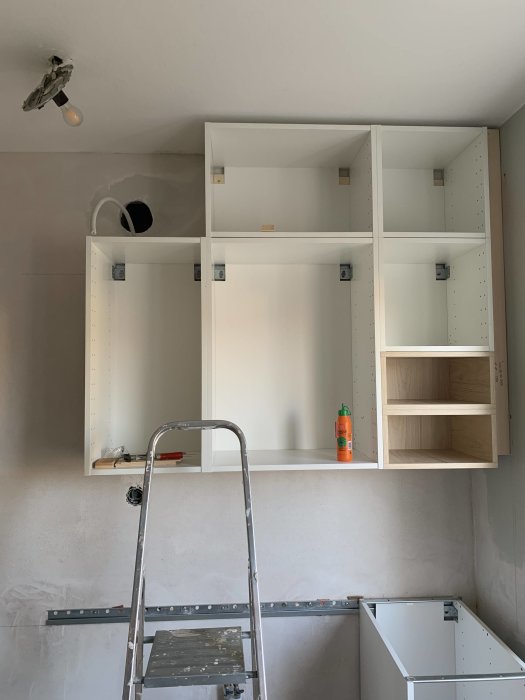 Köksskåp monterade på vägg med uttag för ventilation och kontaktuttag synliga, stege i förgrunden.